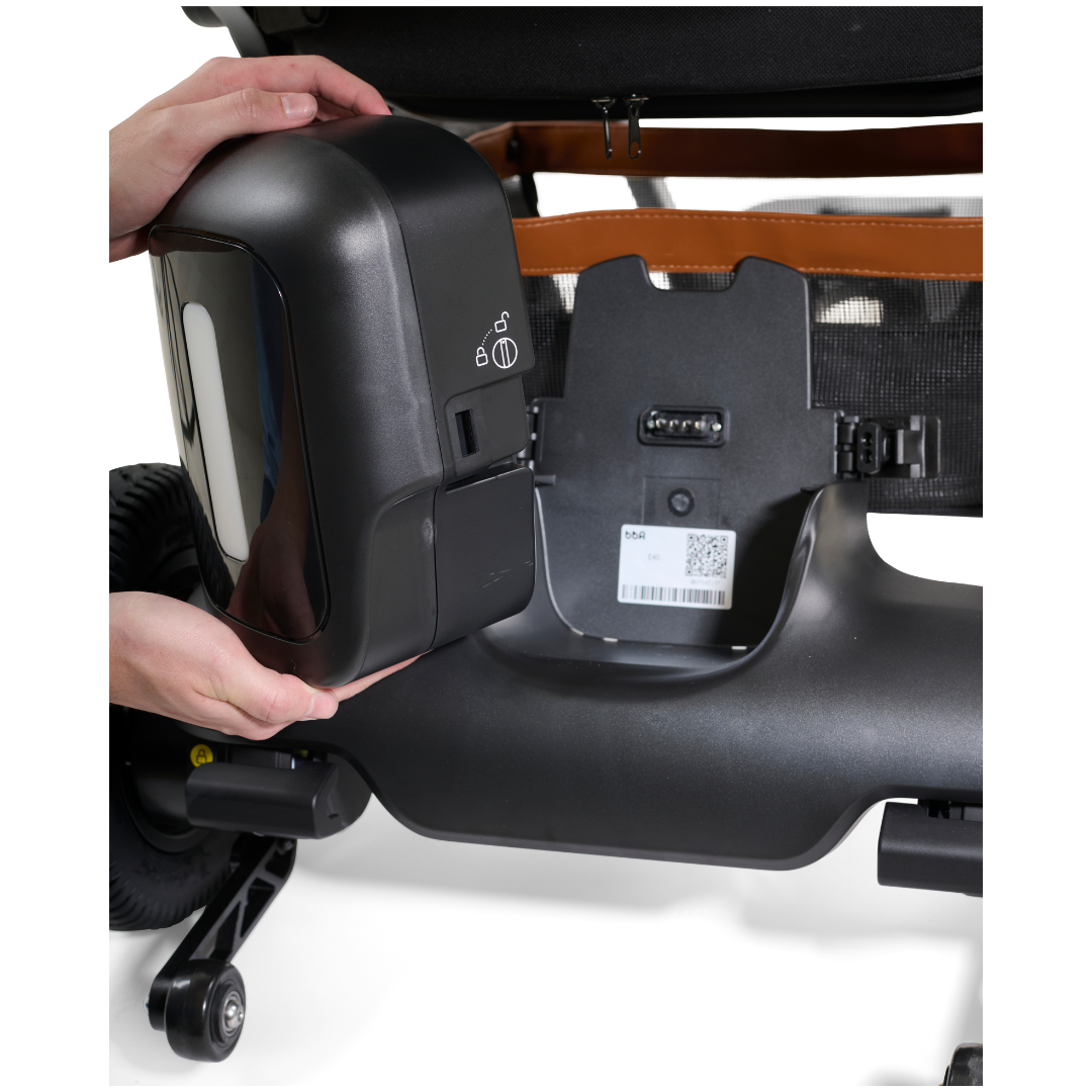 A person carefully removes a rectangular black battery with a QR code and warning symbols from an electric wheelchair, likely to install Golden Tech Accessories for the Ally Powerchair. The metallic frame and wheels exude a wireless remotes functional elegance.